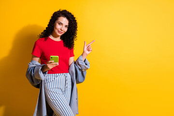 Photo of girl hold phone indicate finger empty space look camera wear striped jeans red t-shirt jacket isolated yellow color background