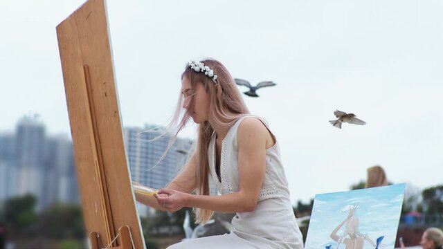 Young blond woman paints a picture on the beach. On open air. Against the background of flying seagulls.