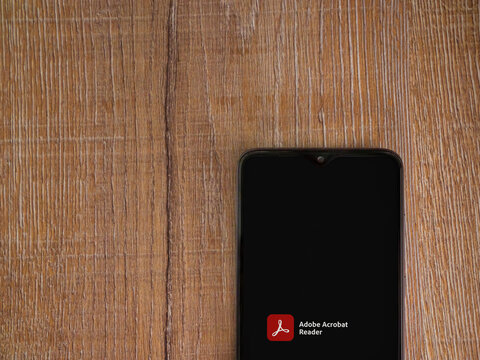 Lod, Israel - July 8, 2020: Adobe Acrobat Reader app launch screen with logo on the display of a black mobile smartphone on wooden background. Top view flat lay with copy space.