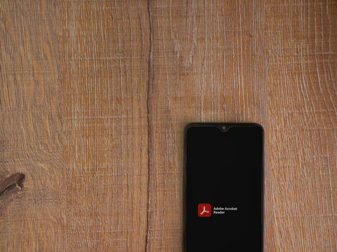 Lod, Israel - July 8, 2020: Adobe Acrobat Reader app launch screen with logo on the display of a black mobile smartphone on wooden background. Top view flat lay with copy space.
