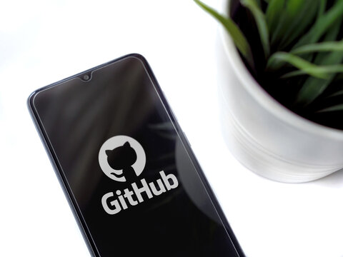 Lod, Israel - July 8, 2020: Modern minimalist office workspace with black mobile smartphone with GitHub app launch screen with logo on white background. Top view flat lay with copy space.