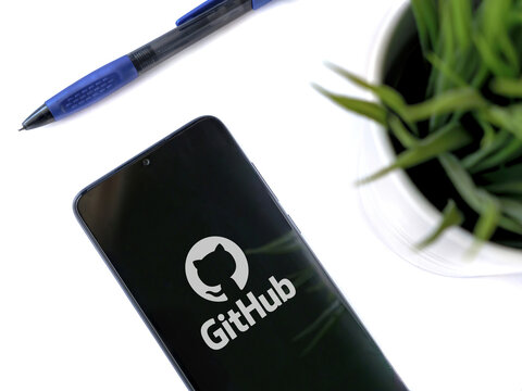 Lod, Israel - July 8, 2020: Modern minimalist office workspace with black mobile smartphone with GitHub app launch screen with logo on white background. Top view flat lay with copy space.