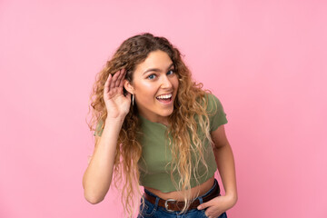 Young blonde woman with curly hair isolated on pink background listening to something by putting hand on the ear