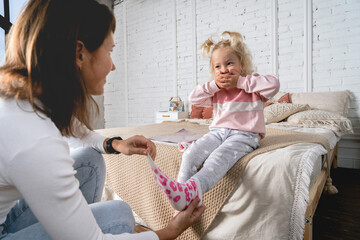 Mom takes off her little daughter's socks and tickles her feet a little. The girl laughs. - obrazy, fototapety, plakaty
