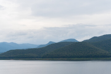 Landscape of the lake dam in the north of Thailand, fertile green forest trees mountain and white blue clouds winter sky as the abstract background, still and calm water