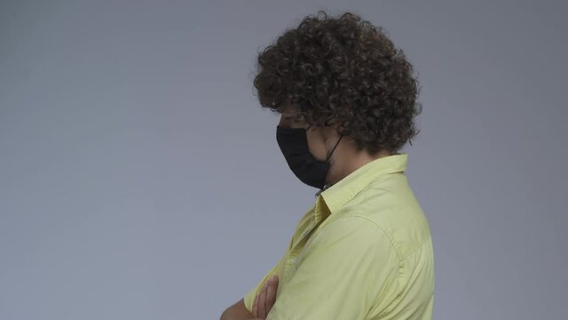 Curly man stands in a profile in a medical mask. Guy crossed his arms on his chest and turns discontentedly into the camera. High quality.