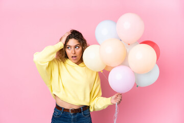 Fototapeta na wymiar Young blonde woman with curly hair catching many balloons isolated on pink background with surprise facial expression