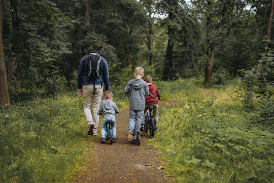 A young caucasian man and three cute boys walking along a path in forest in summer time. One boy is cycling by bicycle, second by runbike, third is riding a kick scooter
