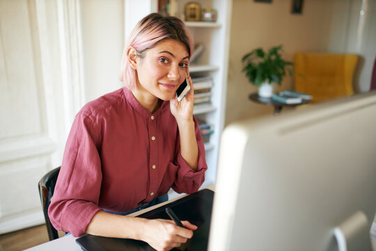 Stylish skilled young female graphic designer with pinkish hair sitting at her workplace using computer and tablet, sketching, making web design, talking on cell phone. Job and creativity concept