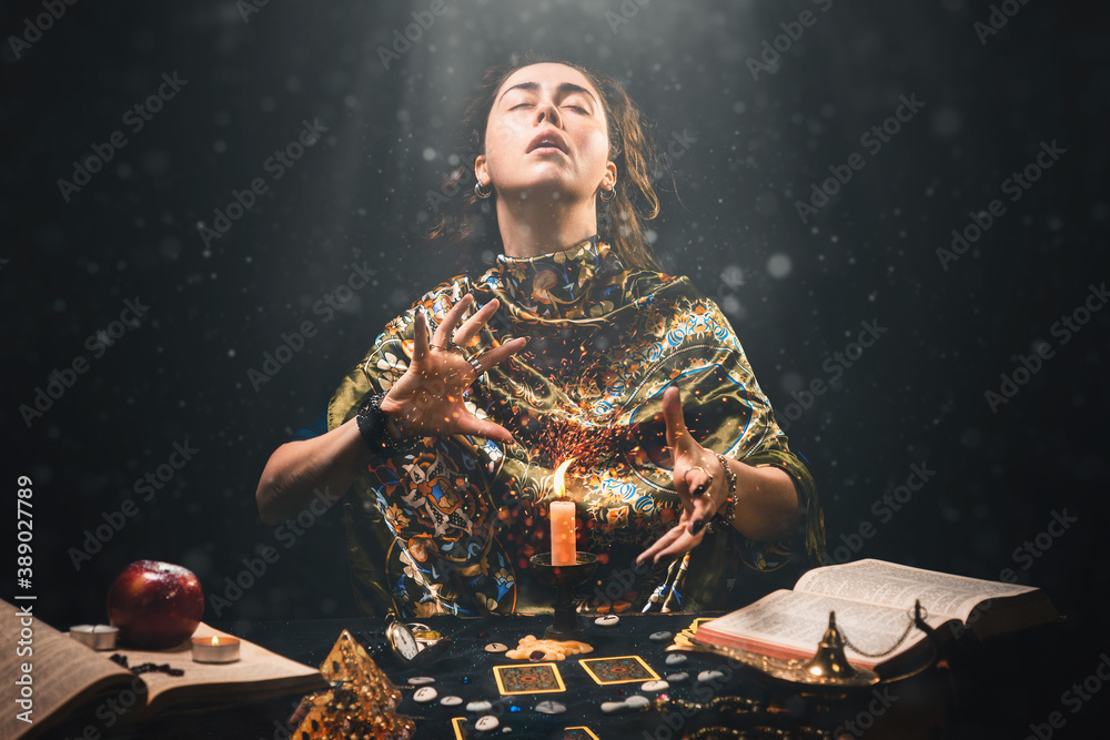 Wall mural Astrology and divination. Portrait of a fortune teller who reads by candle, eyes closed in ecstasy. Black background. There are magic items on the table