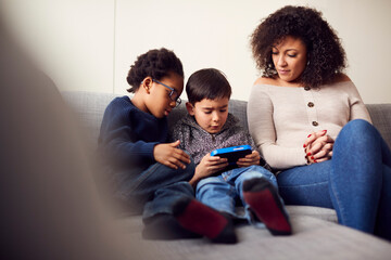 Mother And Sons Sitting On Sofa At Home Playing Computer Game Together On Hand Held Device At Home