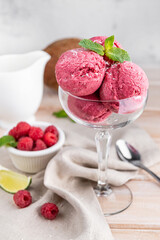 Balls of raspberry ice cream in a transparent glass, decorated with coconut and mint leaves on a light wooden background with raspberries and a slice of lime.