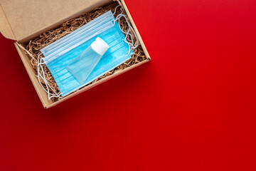 Packing a Christmas present during Coronavirus epidemic. Face masks and hydroalcoholic gel inside a cardboard box on a red background.