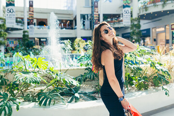 Cheerful lady in stylish black dress carrying on colorful shopping bags while walking at store. Attractive brunette looks satisfied after shopping at mall.