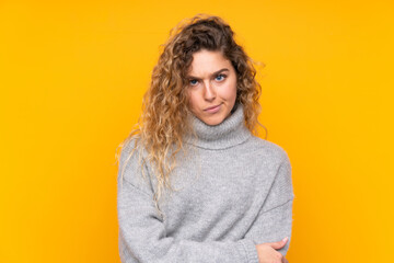 Young blonde woman with curly hair wearing a turtleneck sweater isolated on yellow background feeling upset