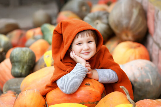 A small/ little child girl in an orange cloak / poncho with a dwarf long hood on a pile / slide of colorful pumpkins. Bright autumn picture,  