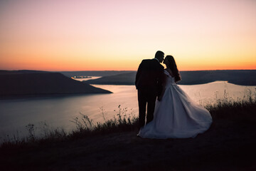 Silhouettes of bride and groom in wedding dress at night against backdrop of large lake and islands. Bakota, Ukraine