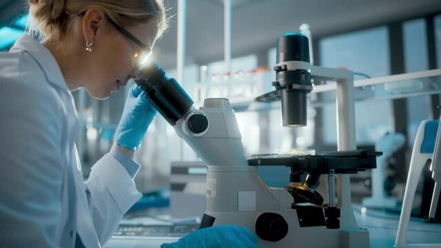In Bright Medical Science Laboratory: Beautiful Microbiologist Looks Under Microscope Does Analysis of Test Sample. Brilliant Scientist, Microbiologist, working with High-Tech Equipment. Close-up