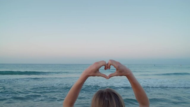 Woman make heart sign with her fingers. Focused on beach and horizon. Concept love for nature and consciousness about ecological projects. Romantic getaway vacation vibes