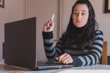 Una mujer joven usa un ordenador portátil para realizar compras por internet mientras sostiene una tarjeta de crédito - obrazy, fototapety, plakaty