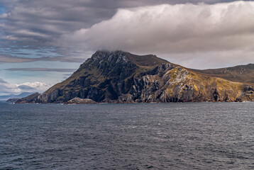 Cape Horn, Wollaston Islands, Chile - December 14, 2008: Dark rock with white and yellow patches mountain rises sharply out of  dark gray ocean and put its top in cloudscape with light blue patches.