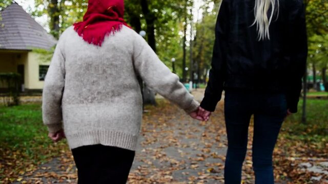 Grandmother and granddaughter walk by the hand in the autumn park. Autumn walk of an old grandmother with an adult granddaughter by the hand background. Grandmother and granddaughter walk by the hand