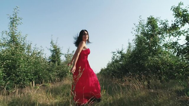 A beautiful Caucasian girl in a red dress runs along the green garden. Dress Folds Out Of The Wind