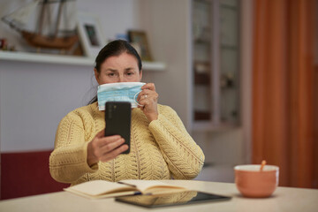 Senior woman in yellow sweater looking at smartphone with mask