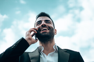 Handsome businessman talking on the cell phone. Bottom up view of a young man smiling in suite talking on the smartphone against a blue cloudy sky