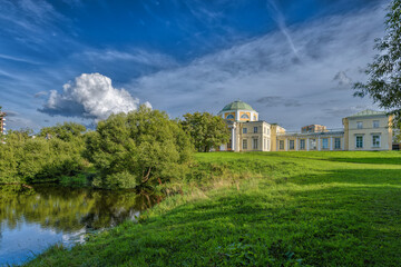 Fototapeta na wymiar The old Palace stands on a hill in front of a pond with a green meadow surrounded by trees against a blue sky with incredible white clouds. Saint-Petersburg, Russia