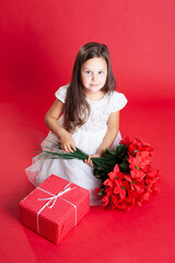 a child in a white dress smiles, holding a gift box and mistletoe in his hands on a red background.