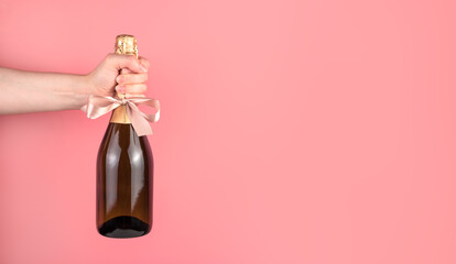Hand holding a bottle of alcohol, decorated with a bow on a pink background. Panorama with space to...