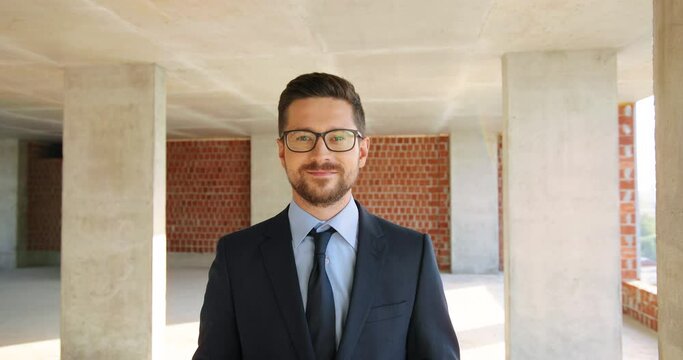 Portrait of Caucasian handsome young man in glasses, suit and tie standing at construction site indoor. Male real-estate agent with tablet device in hands. Zooming in. Dolly shot. Businessman.