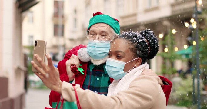 Close up portrait of African American happy female in mask taking selfie photo on smartphone with Caucasian Santa Claus holds bag with presents. Woman recording vlog with Santa on street. X-mas spirit