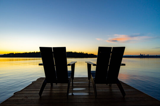 Two chairs at end of dock facing sunset and city in background