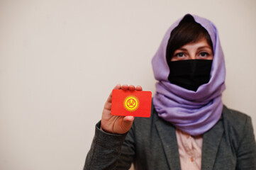 Portrait of young muslim woman wearing formal wear, protect face mask and hijab head scarf, hold Kyrgyzstan flag card against isolated background. Coronavirus country concept.