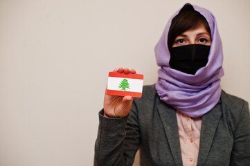 Portrait of young muslim woman wearing formal wear, protect face mask and hijab head scarf, hold Lebanon flag card against isolated background. Coronavirus country concept.