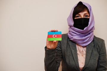 Portrait of young muslim woman wearing formal wear, protect face mask and hijab head scarf, hold Azerbaijan flag card against isolated background. Coronavirus country concept.