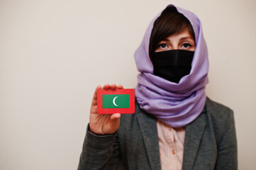 Portrait of young muslim woman wearing formal wear, protect face mask and hijab head scarf, hold Maldives flag card against isolated background. Coronavirus country concept.