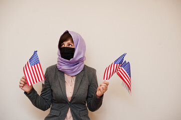 Portrait of young muslim woman wearing formal wear, protect face mask and hijab head scarf, hold USA flags in hands against isolated background. Coronavirus country concept.