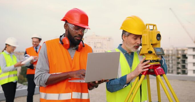 Mixed-races men constructors at building site working and measuring angle with total station on roof. Multi-ethnic males topographers. Caucasian and African American builders with laptop computer.