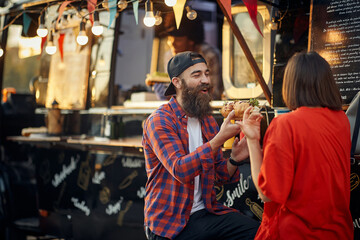 urban couple having fun in front of fast food mobile restaurant, eating one sandwich together, laughing