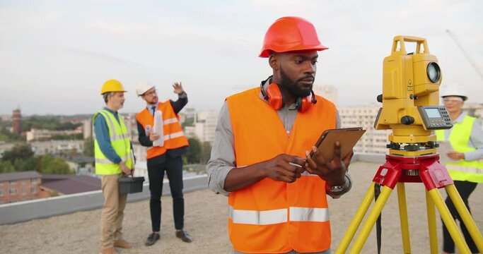 African American man topographer in casque measuring angle with total station on roof of building. Male builder constructor doing topographic measures and using tablet device. Geodesy concept Geodesic