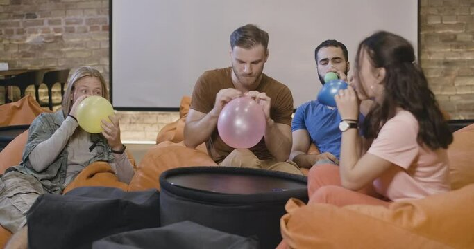 Relaxed multiethnic young friends sitting on bag chairs and blowing balloons. Portrait of cheerful Caucasian and Middle Eastern men and woman having fun. Cinema 4k ProRes HQ.