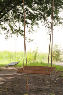 Improvised wooden swing hanging from a tree in the shade, tied up with old rope, during a hot summer day with strong light in the backrgound