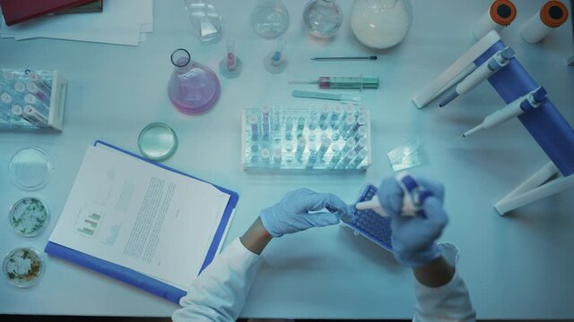 Above the Head Camera Footage of Medical Research Scientists in Blue Rubber Gloves Working Behind Tables in a Modern Laboratory. Doctors Using Test Tubes, Compare Samples and Write Down Data.