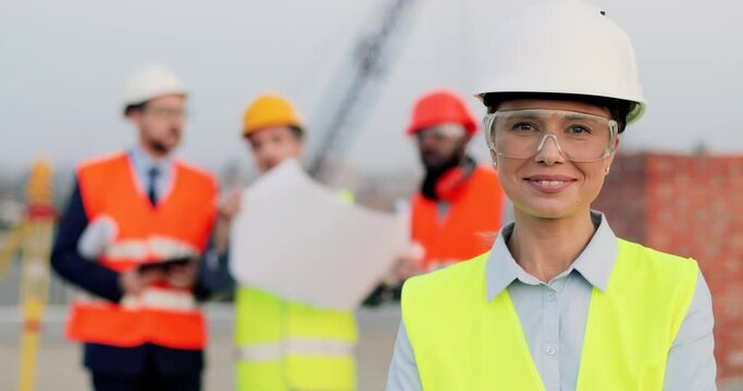 Portrait of beautiful Caucasian young woman in casque and goggles standing at construction site and smiling to camera. Female builder at building. Mixed-races men constructors on background.