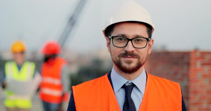 Portrait handsome Caucasian young man in casque and glasses standing at construction site and smiling to camera. Male builder at building. Mixed-races men and woman constructors talking on background.