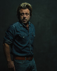 Atmospheric dark studio portrait of a tough middle-aged man with blonde hair in a jeans shirt.