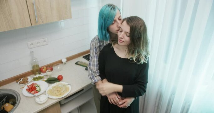 Young lesbian couple hugging in kitchen. High angle of young woman with dyed hair embracing and kissing happy girlfriend on cheek while standing in cozy kitchen during romantic dinner